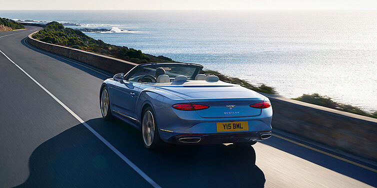 Bentley Tbilisi New Bentley Continental GTC Azure convertible rear view in Light Windsor blue paint with 22 inch ten spoke wheels driving dynamically on an ocean road in the sunshine